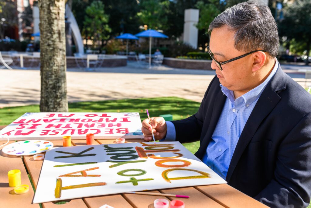 Image of a man with disabilities and of Asian descent with gray hair and glasses painting a sign that reads, ‘Talk to your doctor,’ while at a table outside.